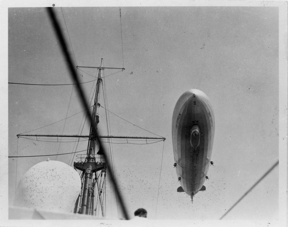 Die historische Aufnahme zeigt die Begegnung eines Zeppelins mit einem Schiff. Unten im Bild ist der Fahnenmast des Schiffs zu sehen, oben fliegt der Zeppelin in der Luft.