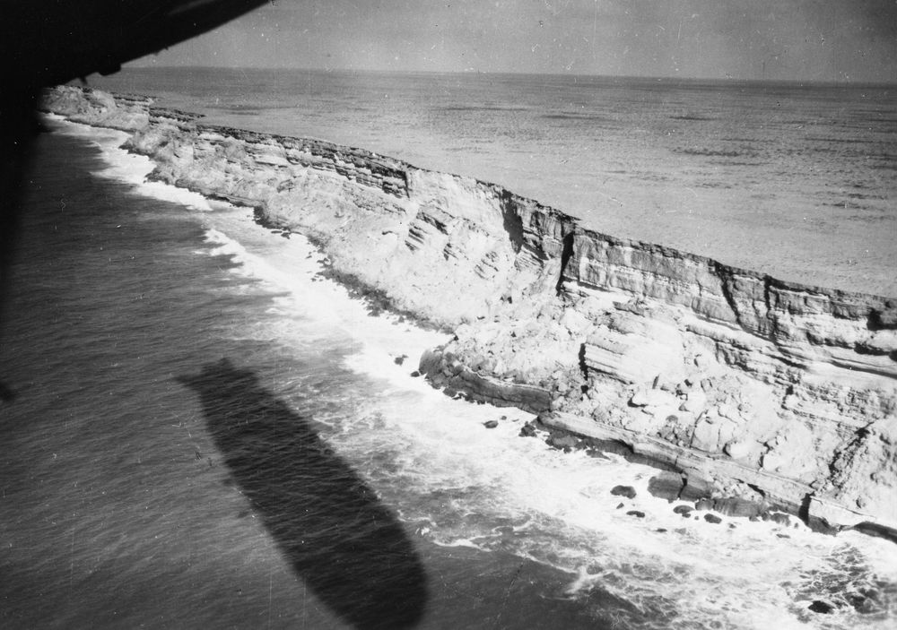 Die historische Aufnahme zeigt neben einer steilen Künste unter der das Meer schäumende Wellen schlägt, den Schatten eines Zeppelins auf der Wasseroberfläche.