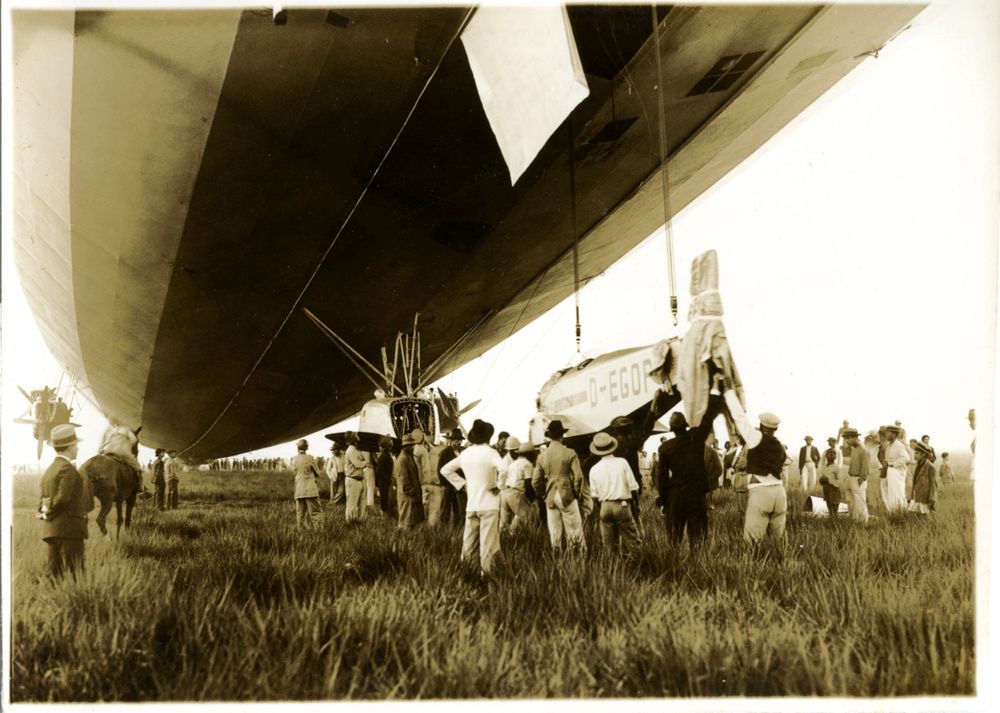 Die historische Aufnahme zeigt das Ausladen eines Flugzeuges aus einem Zeppelin. Um das Geschehen stehen einige Männer.