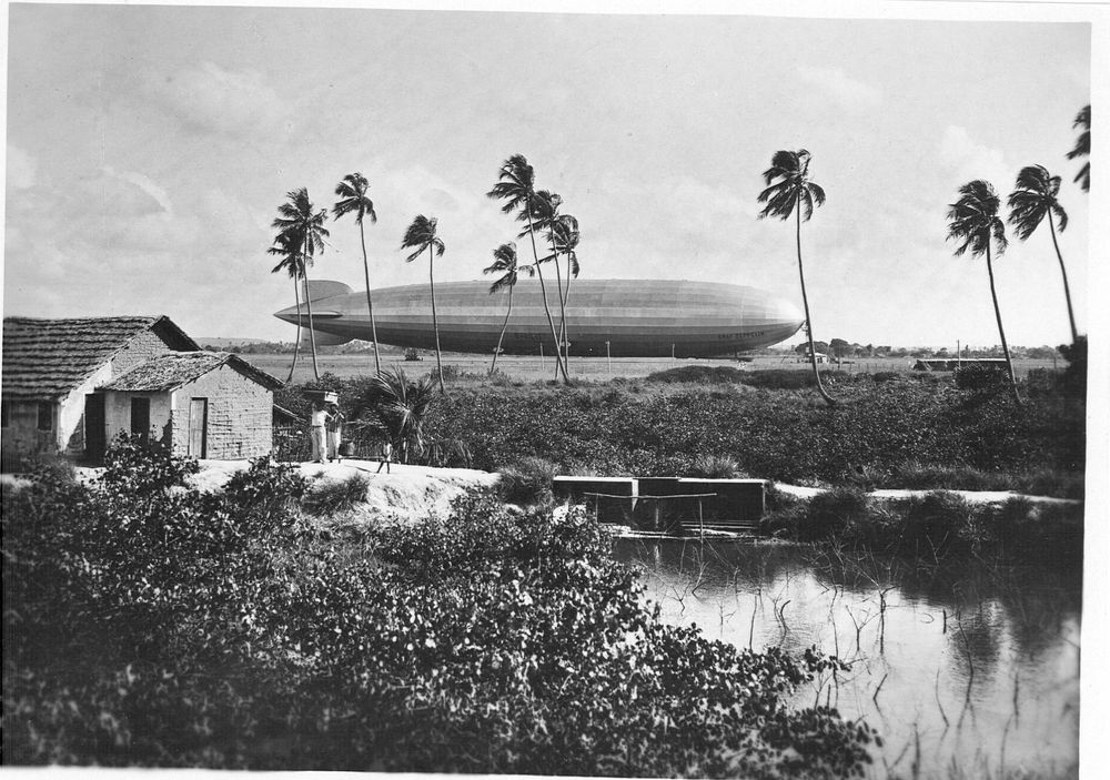 Die historische Aufnahme zeigt einen Zeppelin, der an einem Ankermast befestigt ist. Davor stehen einige Palmen und links im Bild ein Haus.
