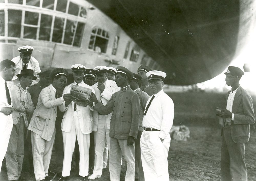 Die historische Aufnahme zeigt eine Gruppe von Männern, die Besatzungsmitglieder des Zeppelins, die eine Zeitung anschauen. Im Hintergrund sieht man die Gondel des Zeppelins. 