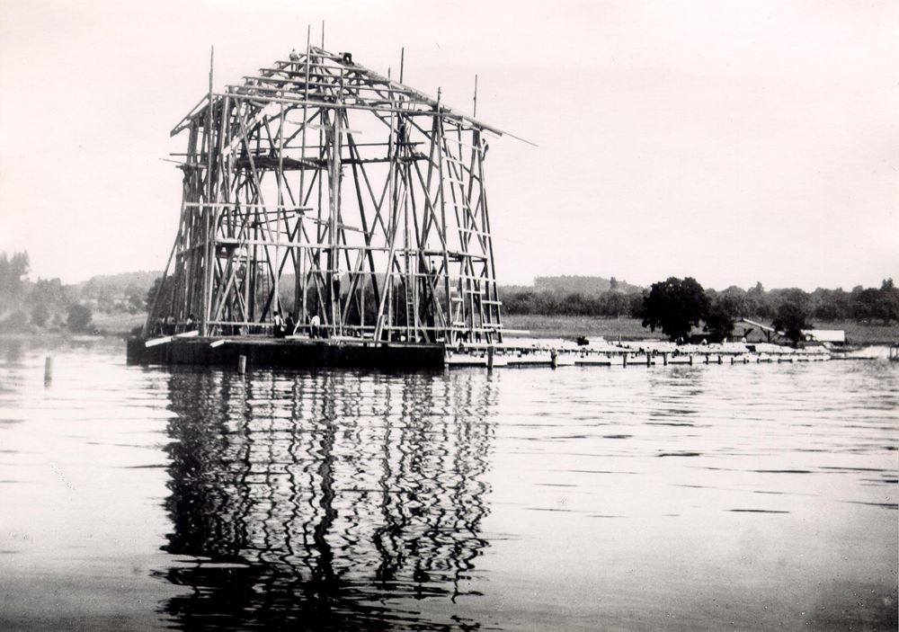 Die historische Aufnahme zeigt die schwimmende Luftschiffhalle während des Baus. Auf dem Bild besteht die Halle erstmal nur aus einem Gerüst aus Holzpfählen.