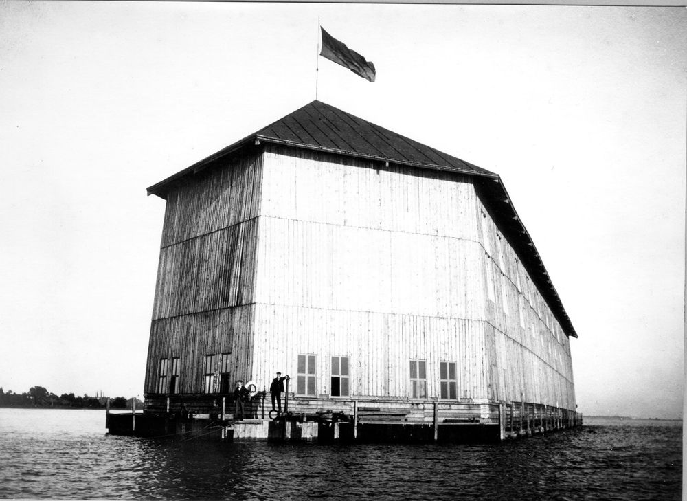 Die historische Aufnahme zeigt die erste schwimmende Halle für Luftschiffe. An einem Steg um die Halle stehen zwei Personen. 