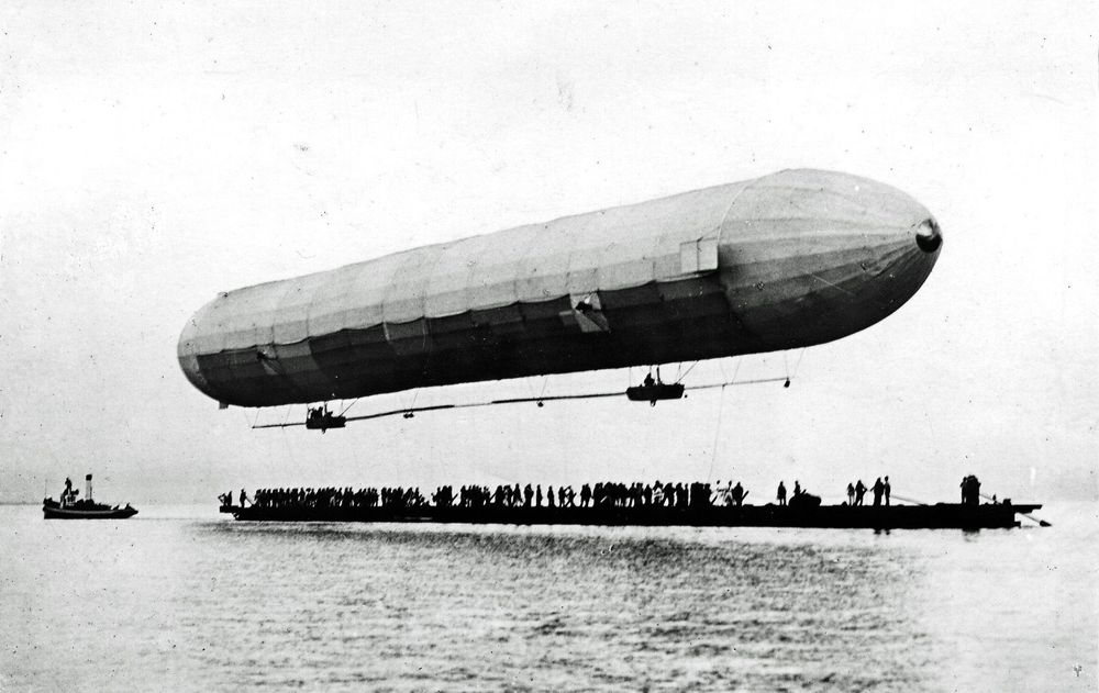 Die historische Aufnahme zeigt ein Luftschiff, das nur einige Meter über dem Wasser schwebt. Dahinter treibt ein sehr langes Boot, auf dem sich viele Personen befinden.