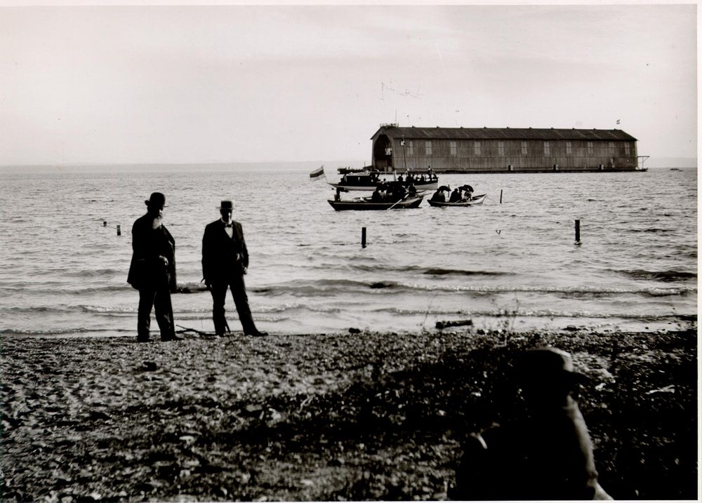 Die historische Aufnahme zeigt zwei ältere Herren, die am Seeufer entlangspazieren. Im Hintergrund auf dem See treibt die eine schwimmende Luftschiffhalle.