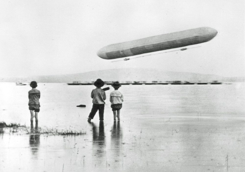 Die historische Aufnahme zeigt drei kleine Kinder, die am Seeufer stehen und einen aufsteigenden Zeppelin betrachten.