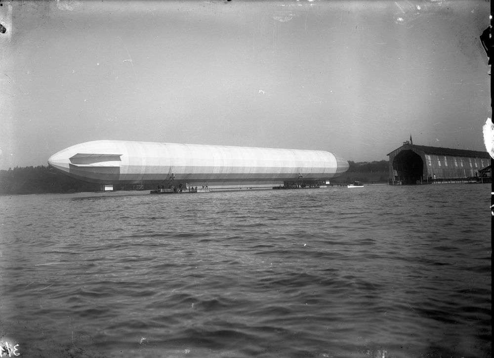 Die historische Aufnahme zeigt ein Luftschiff, das sich auf dem Wasser befindet. Dahinter ist eine Luftschiffhalle zu erkennen.