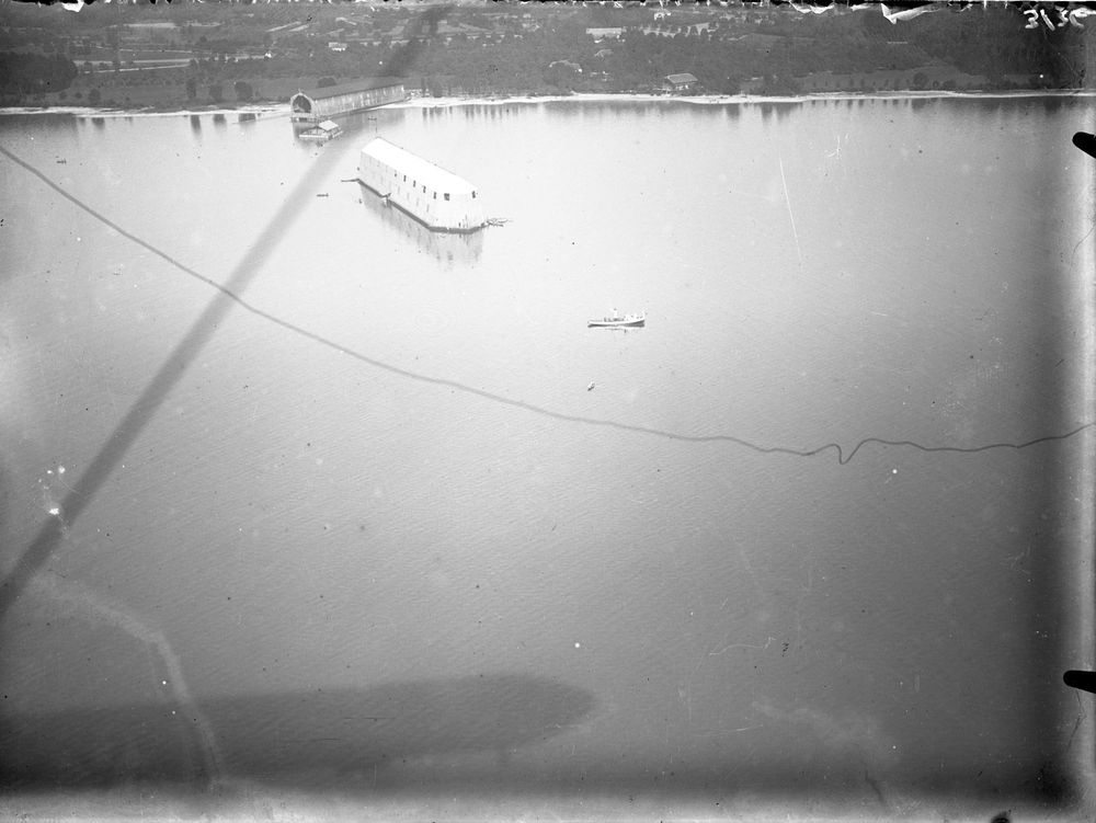 Die historische Aufnahme zeigt eine auf dem Bodensee schwimmende Luftschiffhalle aus der Vogelperspektive. Aufgenommen von einem Zeppelin aus, dessen Schatten auf der Wasseroberfläche zu erkennen ist.