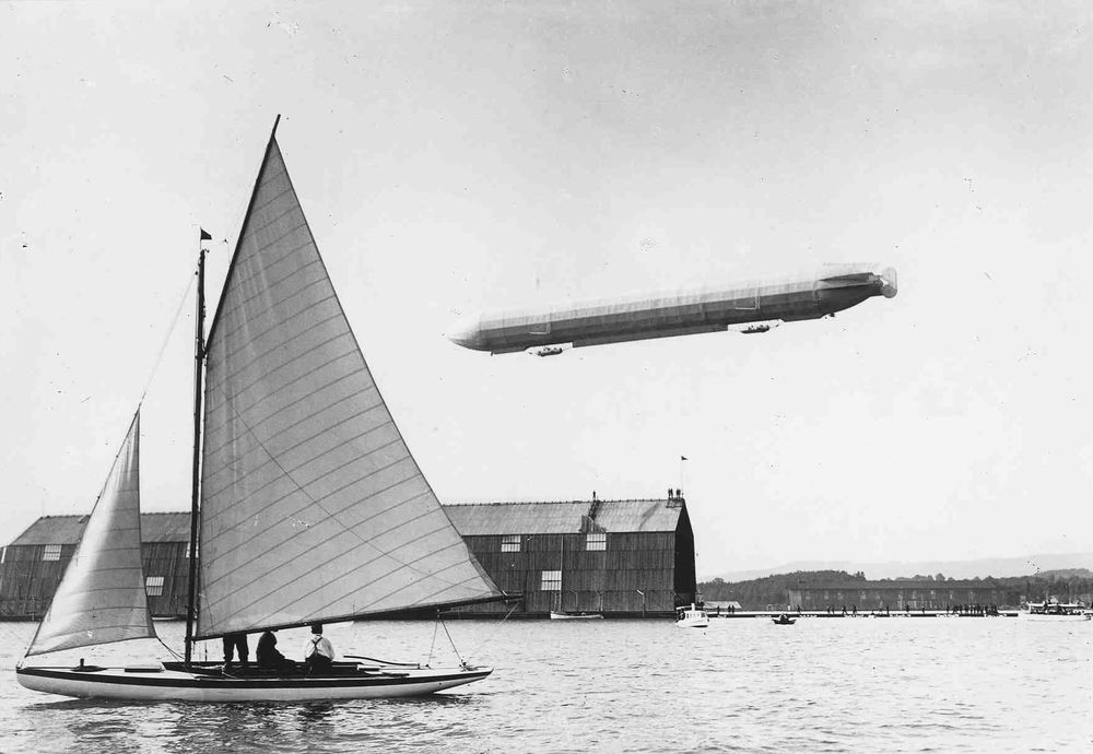 Die historische Aufnahme zeigt eine Luftschiffhalle am See über der ein Zeppelin schwebt. Die Halle wird von einem Segelboot, welches im Vordergrund auf dem Wasser treibt, verdeckt.