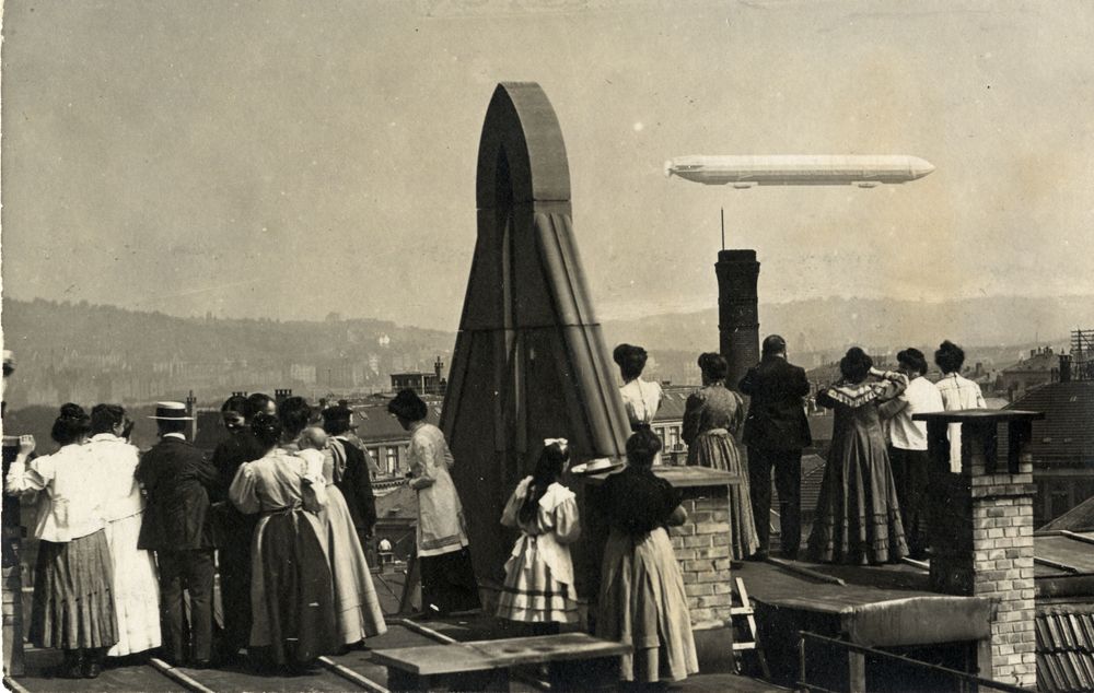 Die historische Aufnahme zeigt eine Gruppe von Menschen, die auf einem Hausdach steht und in der Ferne einen vorbei fliegenden Zeppelin beobachtet.