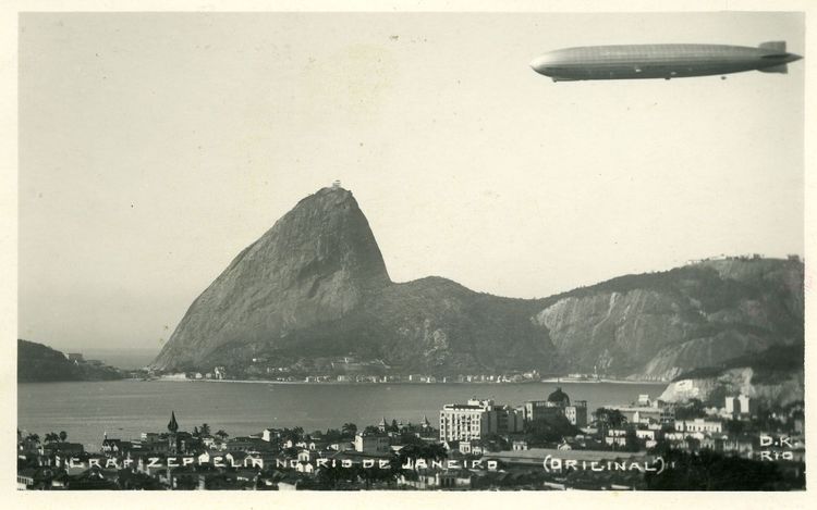 Die historische Aufnahme zeigt einen Zeppelin der über Rio de Janeiro fliegt. Unterhalb sieht man die Stadt und das angrenzende Meer und dahinter große Berge. Darunter der Zuckerhut am Eingang zur Guanabara-Bucht.