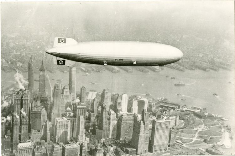 Die „Hindenburg“ über den Hochhäusern von New York. Das Luftschiff ist zentral im Bild, im Hintergrund ragen Piers ins Wasser.