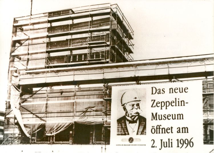 Schwarz-Weiß-Foto des Hafenbahnhofs im Umbau. Das Gebäude ist mit einem Gerüst mit Planen umstellt. Ein großes Plakat zeigt Graf Zeppelin mit einem Bauarbeiterhelm zu sehen. Rechts vom Portrait steht: Das neue Zeppelin-Museum eröffnet am 2. Juli 1996.